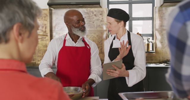 Diverse group of chefs working together in a professional modern kitchen. Two chefs engaged in a conversation, one holding a mixing bowl and the other holding a tablet or recipe book. Ideal for illustrating culinary training, teamwork, and professional cooking environments. It can be used for culinary school brochures, teamwork and leadership articles, cooking blogs, and restaurant promotional materials.