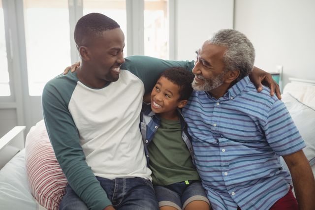 Three generations of a family sitting together on a bed, smiling and bonding. The grandfather, father, and son are sharing a moment of happiness and togetherness. This image can be used for family-oriented content, advertisements promoting family values, or articles about multi-generational relationships.