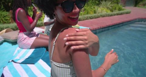 African american women applying sunscreen and laughing poolside on striped towel in backyard