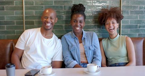 African friends laughing and sitting in cozy cafe booth with coffee cups and smartphone