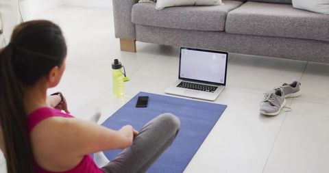 Korean woman stretching on blue yoga mat during guided home workout with laptop and sneakers