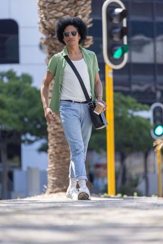 Sunlit urban commute man walking with coffee and crossbody bag wearing sunglasses and jeans