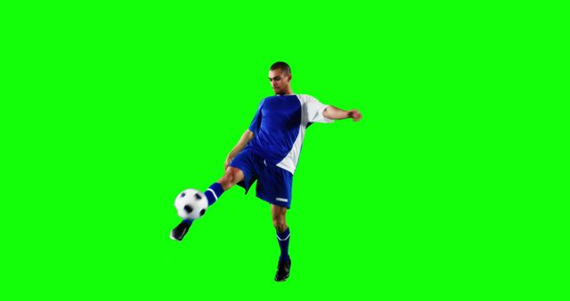 A young Caucasian male athlete in a blue soccer uniform is kicking a soccer ball mid-air against a green screen background, with copy space. His dynamic pose captures the action and skill involved in the sport of soccer.