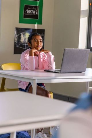 Asian schoolgirl sitting at desk using laptop in bright classroom wearing pink hoodie