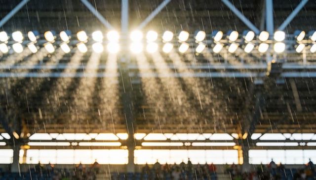 Backlit stadium rain streaming through floodlights over silhouetted crowd under steel trusses