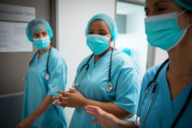 Image shows a group of healthcare professionals, including both male and female doctors, walking in a hospital corridor while wearing face masks and blue scrubs. They appear to be engaged in a discussion, indicating teamwork and collaboration during the COVID-19 pandemic. Useful for depicting healthcare settings, medical discussions, teamwork in healthcare, and protective measures during the pandemic.