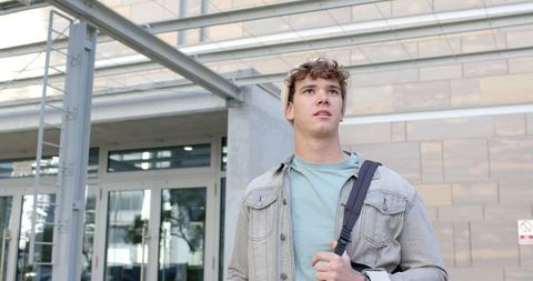 Young commuter standing outside modern campus building carrying backpack and looking ahead