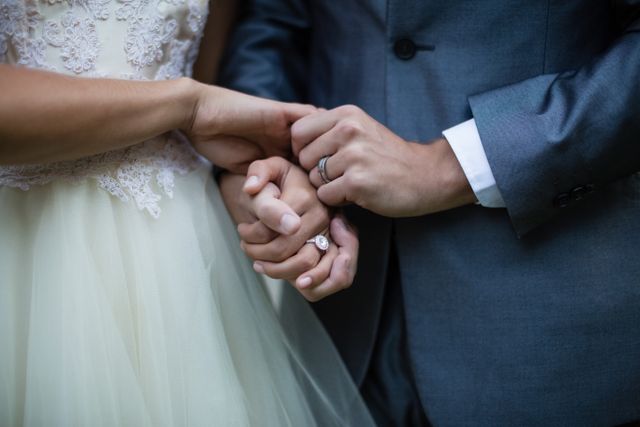 Close-up view of a wedding couple holding hands, showcasing their wedding rings. The bride is wearing a lace wedding dress, and the groom is in a grey suit. Perfect for use in wedding invitations, romantic greeting cards, wedding blogs, and articles about marriage and commitment.
