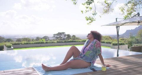 African american woman relaxing by infinity pool on luxury terrace wearing floral cover-up