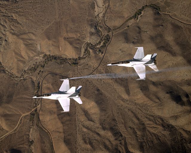 F/A-18 fighter jets executing precision formation flight over barren, rugged terrain. Smoke trails highlight the wingtip vortices induced by the aircraft, creating an aerodynamic visual pattern. Useful for topics on military aviation, flight formation techniques, air performance, and aerodynamics.