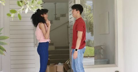 Diverse couple chatting on front porch while checking smartwatch and unpacking boxes