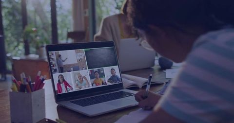 Girl writing while joining virtual classroom on laptop at home with pencil cup and notebook