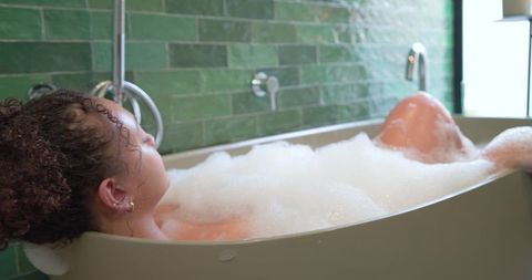 Woman reclining in foam bath in freestanding tub with green glazed tile and natural light