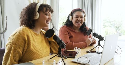 Female podcasters recording, laughing in home studio with laptop, headphones, microphones