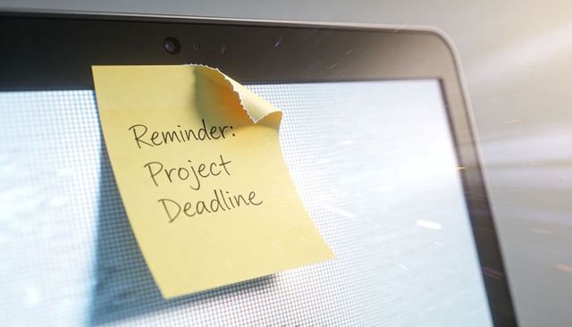 Yellow sticky note reminding of project deadline on laptop screen with webcam glare