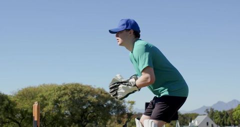 Crouching wicketkeeper wearing blue cap and gloves ready behind stumps sunny cricket field