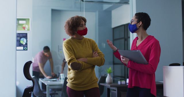 Two colleagues are seen discussing a project while wearing face masks in a modern office. They stand facing each other, demonstrating active communication and collaboration. One holds a tablet, implying a discussion of details or plans. In the background, another employee is working at a desk, indicating a busy and productive office atmosphere. This image is ideal for illustrating concepts of modern workplace dynamics, safety measures in businesses during a pandemic, and professional collaboration in a corporate setting.