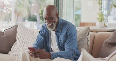 Senior african-american man smiling while using smartphone on cozy living room sofa
