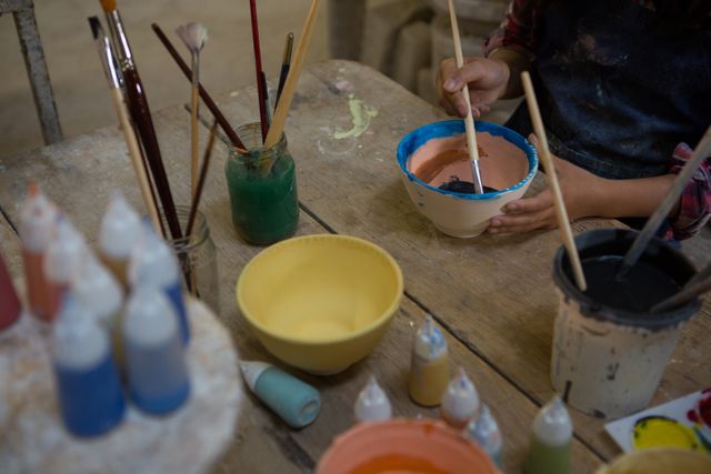 Child painting bowl in pottery workshop. Focus on hands and creative process with paints and brushes on wooden table. Suitable for illustrating art activities, children's hobbies, creative education, handmade crafts, and artistic workshops.
