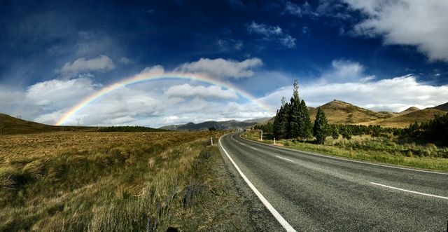 Scenic road stretching through rolling hills with a beautiful rainbow arching across the sky. Ideal for use in travel advertisements, landscaper designs, or outdoor adventure promotions. Illustrates the tranquility and beauty of nature, perfect for inspiration and wanderlust themes.