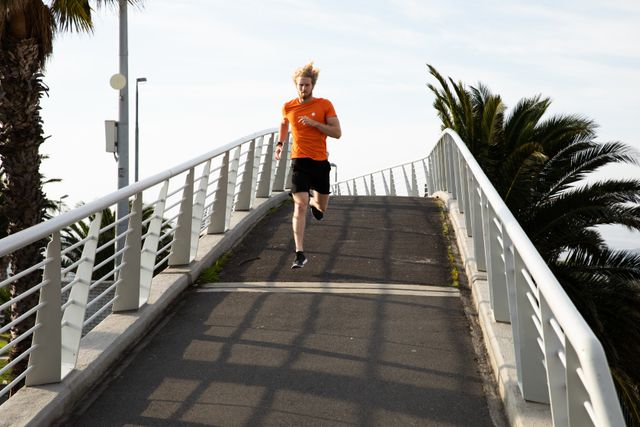 Front view of a fit Caucasian man with long blonde hair wearing sportswear exercising outdoors in the city on a sunny day with blue sky, running on a footbridge.