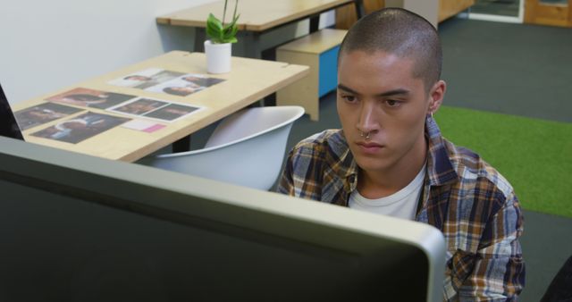 Young man working on computer in a modern office, showing concentration and seriousness. Useful for illustrating productivity, modern workplace, technology usage, student life, and professional environments.