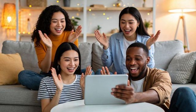 Friends cheering over tablet while clapping and smiling during cozy living room gathering