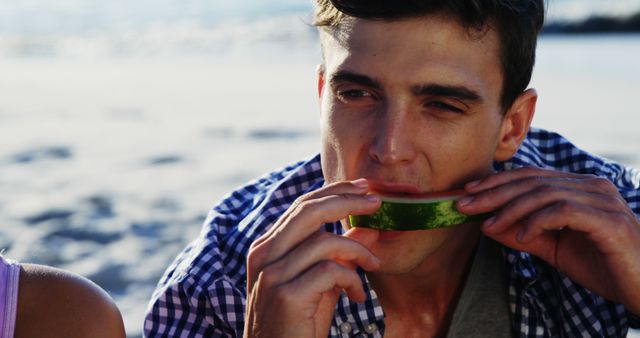 Man enjoying a fresh watermelon slice on a sunny day outdoor, likely at a beach or a picnic spot. He is dressed in a casual striped shirt and appears relaxed. The imagery evokes feelings of summer leisure, relaxation, and healthy living, making it suitable for advertisements related to summer holidays, healthy eating, outdoor activities, and lifestyle promotions.