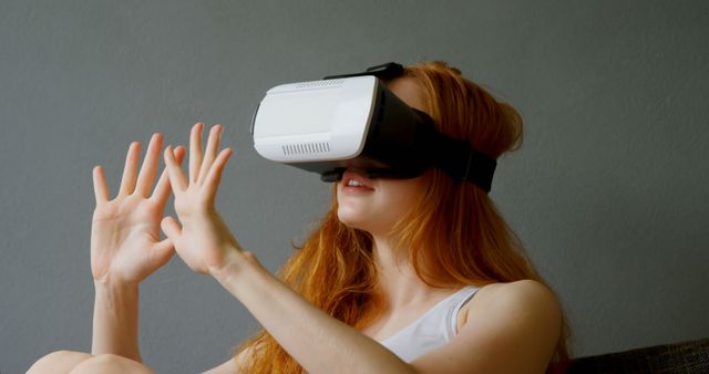Red-haired woman is engaged with a virtual reality headset, utilizing it for an immersive interactive experience. She appears to be engaging with a digital environment, suggesting the widespread use of VR in gaming, entertainment, and educational contexts. This content is ideal for illustrating advancements in consumer tech, virtual reality applications, or the integration of digital and physical worlds.