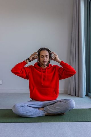 Young man meditating and listening to music with headphones in red hoodie on yoga mat