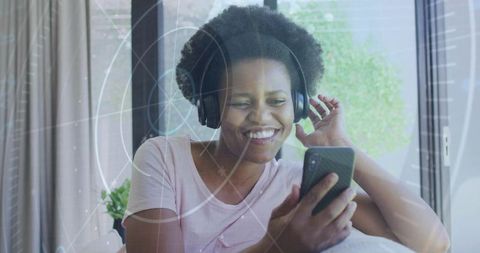 Smiling young woman listening with headphones and using smartphone on bright sofa