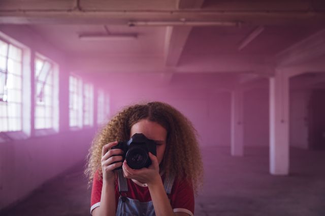 Front view of a hip young Caucasian woman in an empty warehouse, standing and taking photos with SLR camera, wearing a red shirt.