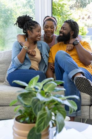 Black family cuddling on sofa in sunlit living room, daughter hugging parents