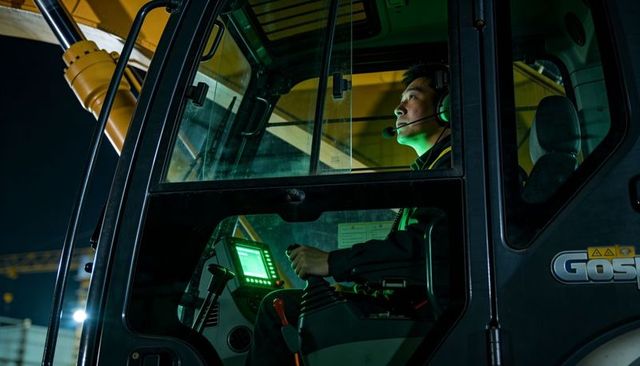 Night crane operator controlling joystick and digital display in illuminated industrial cab