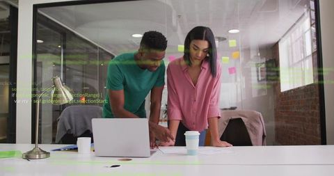 Colleagues leaning over laptop reviewing documents in modern glass-walled office