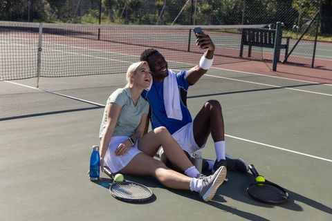 Smiling diverse tennis partners taking selfie on court after match, rackets, water bottles