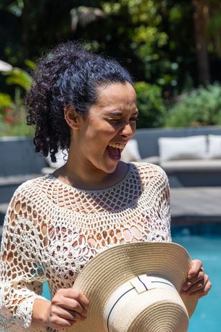African american woman laughing by sunny pool holding straw sunhat wearing crochet coverup