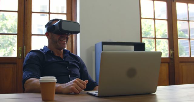 Businessman is using a virtual reality headset while working at his desk in a modern office. The scene captures the intersection of professional work and innovative technology, showing the man engaged with both a laptop and VR. Ideal for use in articles about remote work innovations, the integration of VR in business, or the future of office technology.