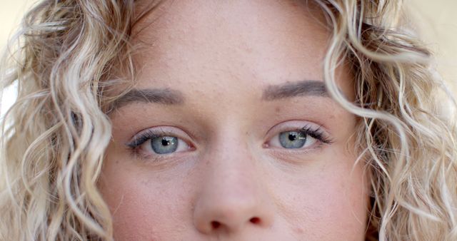 Close-up view of a young woman with curly blonde hair and blue eyes. This can be used to depict themes of beauty, cosmetics, human emotions, or for detailed portraits in artistic or commercial work.