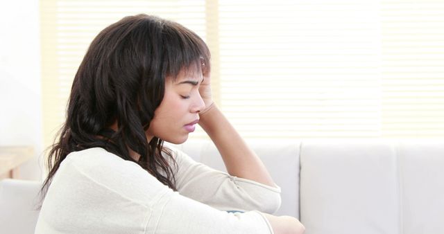 A woman is sitting alone indoors with a hand resting on her head, appearing stressed and reflective. Ideal for use in articles, blogs, and media related to mental health, stress management, emotional well-being, and solitude.