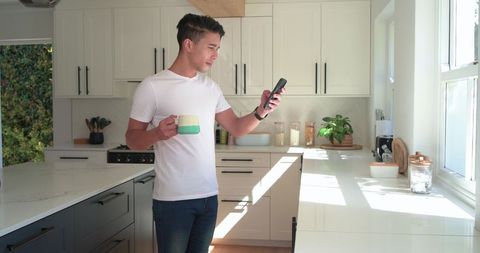African american man standing in modern minimal kitchen holding mug checking smartphone