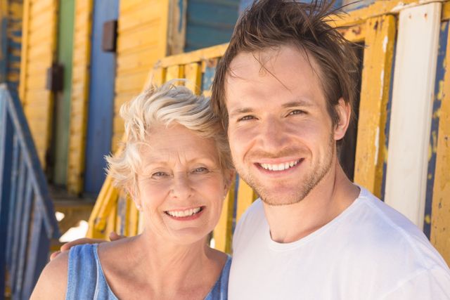 This image captures a joyful moment between a man and his mother standing in front of a vibrant beach hut. Ideal for use in family-oriented content, vacation promotions, or advertisements highlighting family bonding and summer activities.