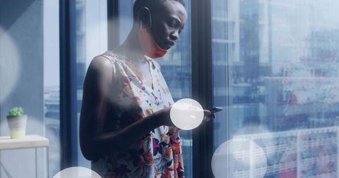 Woman wearing lowered mask checking smartphone by high-rise window with city reflections