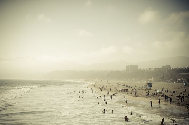 Ideal for depicting a busy summer day at a seaside location, this image captures numerous beachgoers enjoying the waves and sun on a hazy day. Perfect for travel brochures, vacation ads, or lifestyle blogs focusing on coastal getaways and outdoor relaxation.