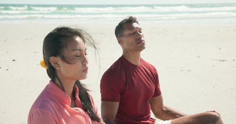 Diverse couple meditating on sunlit sandy beach practicing mindfulness and relaxation