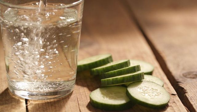 Pouring water into glass with cucumber slices on rustic wood, fresh hydration concept