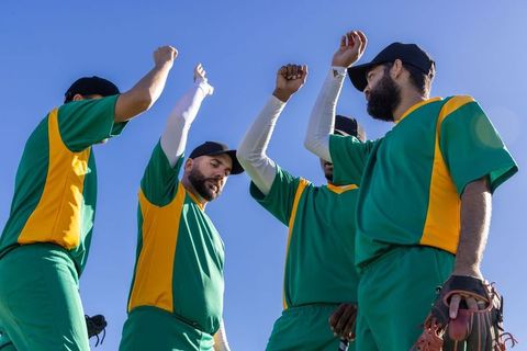 Baseball teammates celebrating high-five in green and yellow uniforms under blue sky