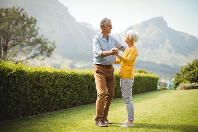 Elderly couple sharing a dance, enjoying a sunny day in a beautiful park with mountains in the background. Ideal for advertisements promoting retirement living, healthy lifestyles for seniors, outdoor activities, and products related to senior wellness.