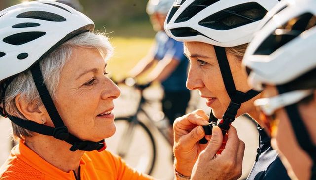 Senior woman adjusting teammate helmet strap during group cycling on sunlit outdoor ride