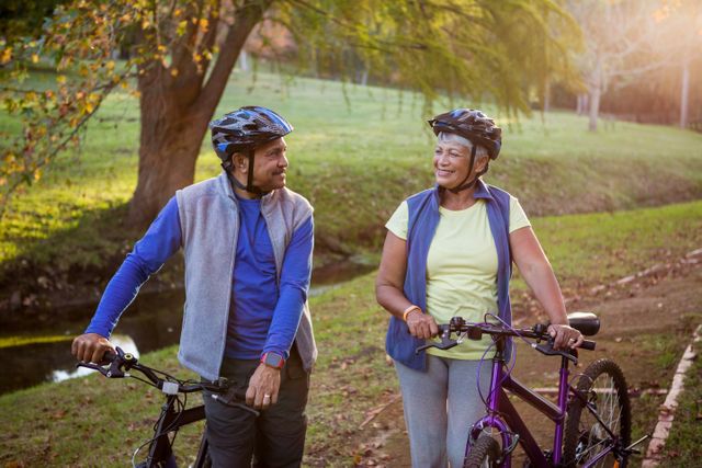 Mature couple enjoying a sunny day in the park while walking their bikes. Both are wearing helmets and casual athletic clothing, indicating a focus on health and fitness. Ideal for use in advertisements promoting active lifestyles, senior fitness programs, outdoor activities, and health and wellness campaigns.